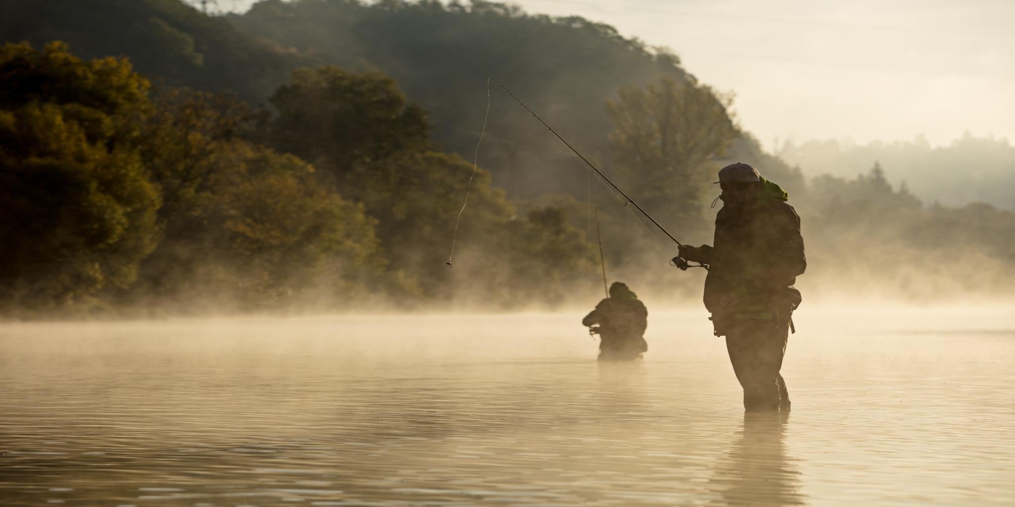 Two anglers wearing waders while fishing in a misty river during sunrise, showcasing the tranquility of nature and the joy of outdoor activities.